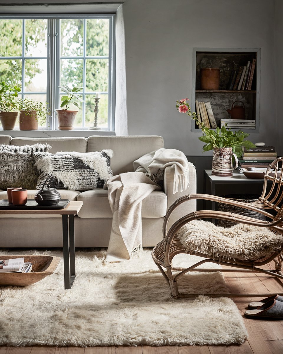 Cozy living room featuring a beige sofa, textured throw blanket, rattan chair with sheepskin, and a plush cream area rug.