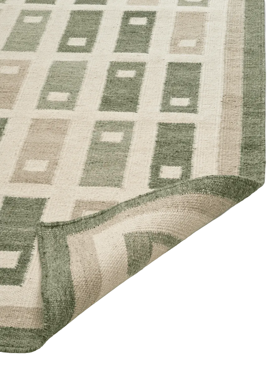 Fyrbo matta i ull med blockmönster i naturnära grönt och beige, flatvävd och vändbar, frilagd produktbild mot ljus bakgrund. Detaljbild av ett uppvikt hörn.