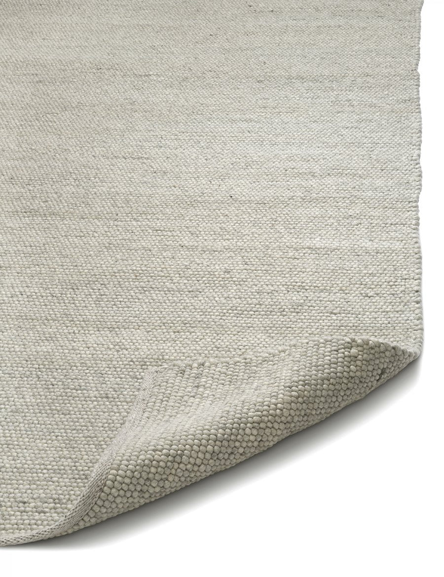 Close-up of a textured, light gray wool area rug with a folded corner, showcasing its chunky woven knit construction.