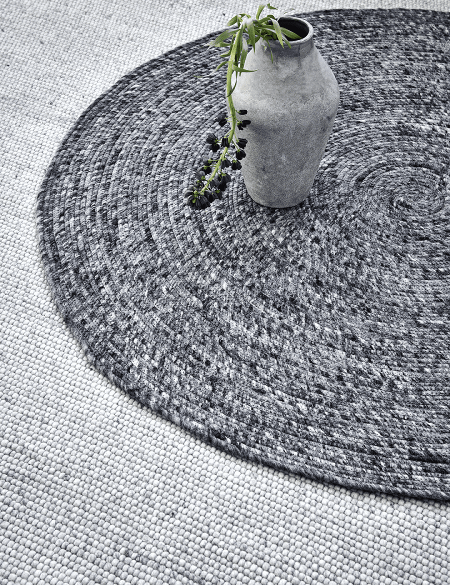 Round gray braided jute rug with a rustic ceramic vase and greenery, set against a neutral textured carpet.