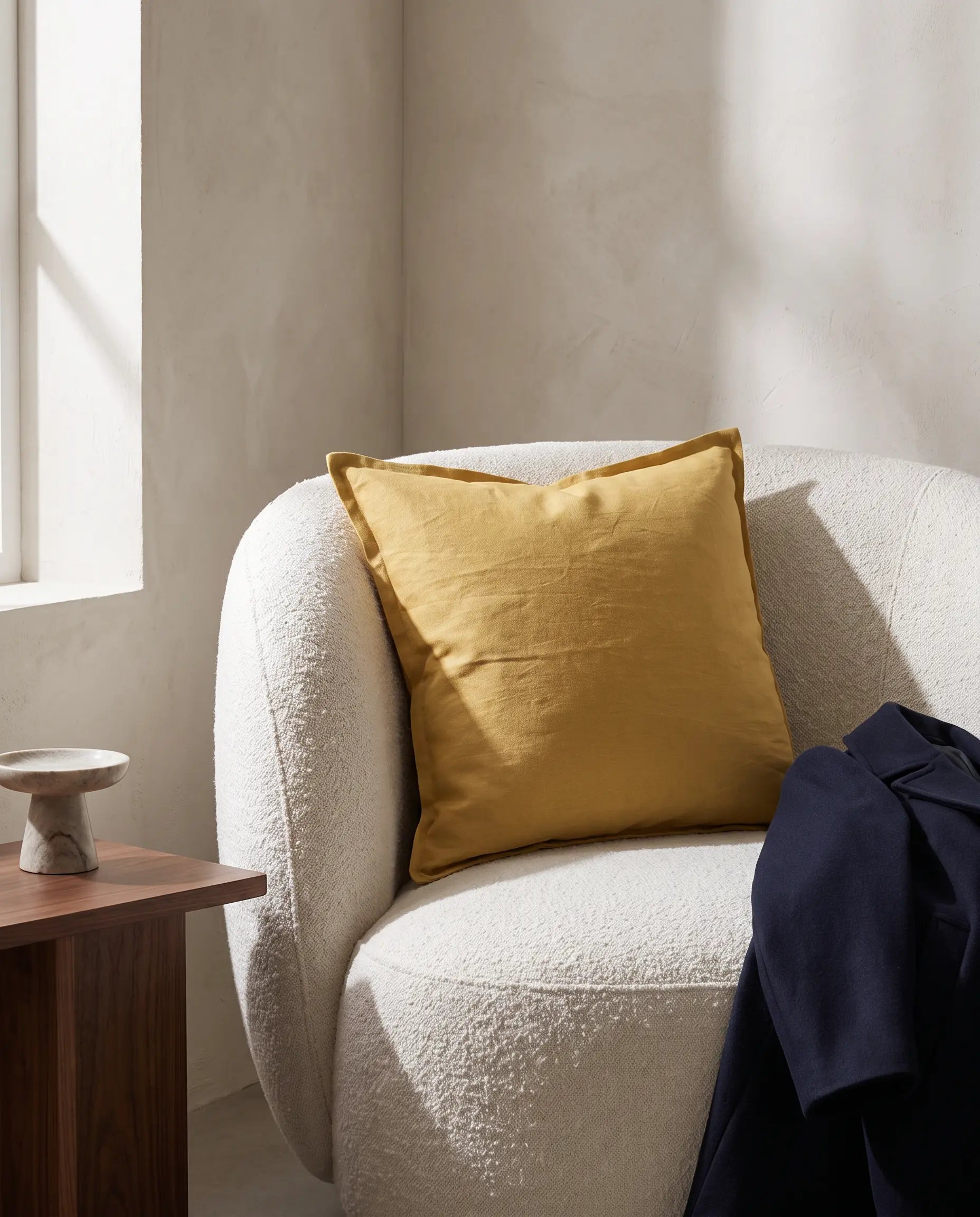 Kuddfodral Classic Linen Ockra 50x50 cm från Classic Collection i enfärgat linne, stylat i en ljus fåtölj i en varm, neutral interiör.