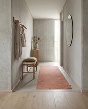 Avlång, terrakottafärgad matta i en minimalistisk hall med betongväggar, träbänk och rund spegel.