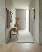 Minimalist hallway featuring a white runner rug, wooden bench, coat rack, and round mirror against modern concrete walls.