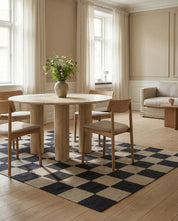 Runder Esstisch aus hellem Travertin mit vier Holzstühlen auf einem schwarz-weiß karierten Teppich in modernem Wohnzimmer.
