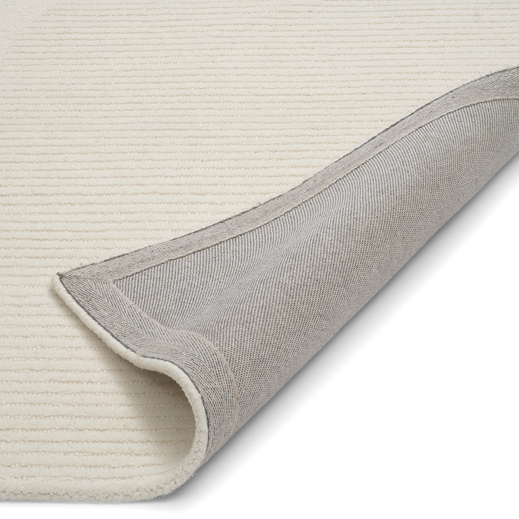 Close-up of a ribbed, cream-colored area rug folded back to reveal the durable gray fabric backing.