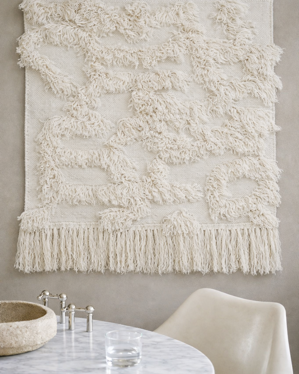 Rio väggbonad i off-white, 100 × 100 cm, handvävd i ull, upphängd på vägg ovanför runt bord i ljus interiör.