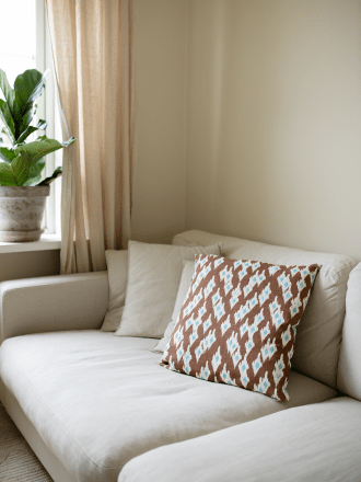 Kuddfodral Ikat Diamond i färgen Mocha, 50x50 cm, placerat i en ljus soffa bredvid ett fönster med linnegardin och grön växt.