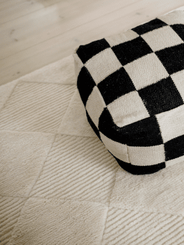 Puff Square Svart/Natur, 50x50 cm, handvävd i ull, placerad på en ljus strukturerad ullmatta.