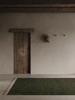 Ullmatta Pure Skogsgrön på betonggolv framför rustik trädörr i dämpad, naturlig miljö.