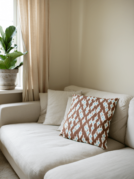 Kissenhülle Ikat Diamond in der Farbe Mocha, 50x50 cm, auf einem hellen Sofa neben einem Fenster mit Leinenvorhang und grüner Pf